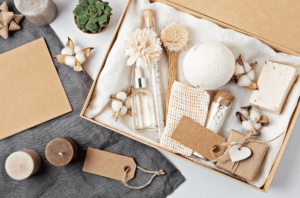 A gift box with bath and body items including a bath bomb, soap, dried flowers, glass bottles, and a scrubber, arranged neatly with brown tags and cotton flowers. Nearby are candles, a plant, and blank cards.