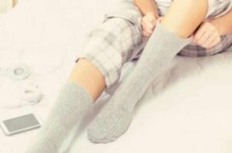 Person sitting on a bed wearing plaid pajamas, pulling up gray socks. A pair of headphones and a smartphone are lying nearby on the bed.