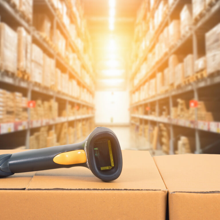 A barcode scanner rests on stacked cardboard boxes in a warehouse with shelves full of inventory management supplies, brightly lit by sunlight in the background.