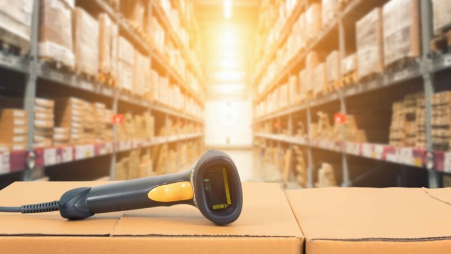 A barcode scanner rests on stacked cardboard boxes in a warehouse with shelves full of inventory management supplies, brightly lit by sunlight in the background.