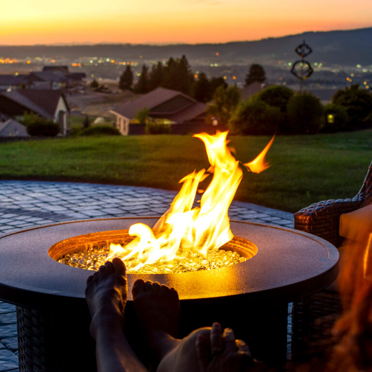 A person relaxes with their feet up near a glowing fire pit on a patio at sunset, overlooking a scenic landscape with houses, grassy hills, and distant city lights.