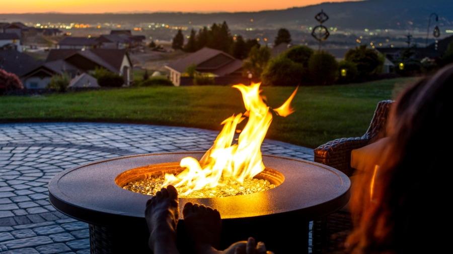 A person relaxes with their feet up near a glowing fire pit on a patio at sunset, overlooking a scenic landscape with houses, grassy hills, and distant city lights.