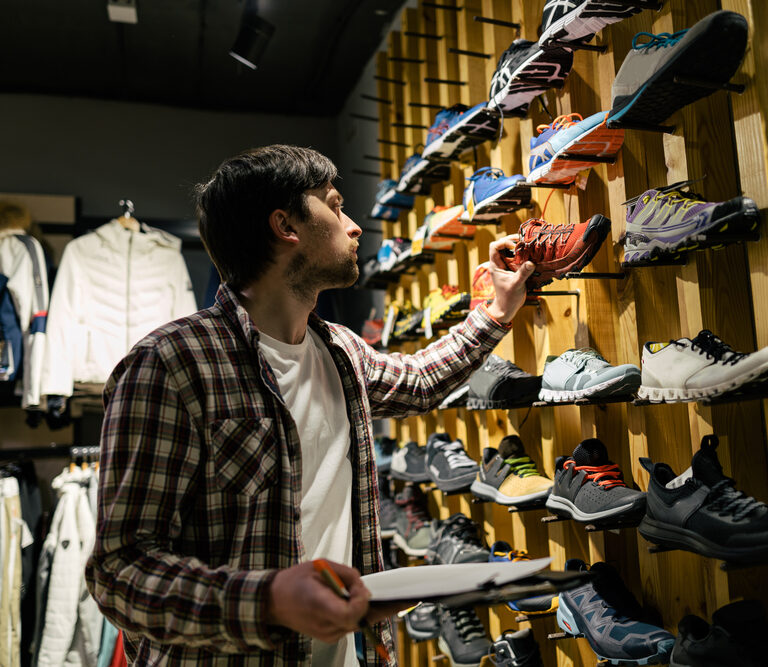 A man in a plaid shirt examines a wall display of athletic shoes in a store, holding a notepad and pen for assortment planning, surrounded by various colorful sneakers and jackets.