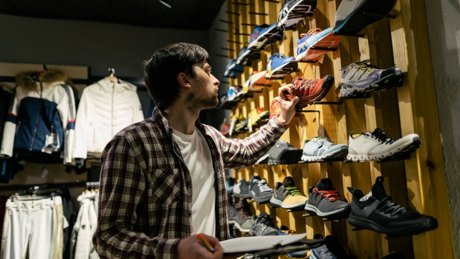 A man in a plaid shirt examines a wall display of athletic shoes in a store, holding a notepad and pen for assortment planning, surrounded by various colorful sneakers and jackets.