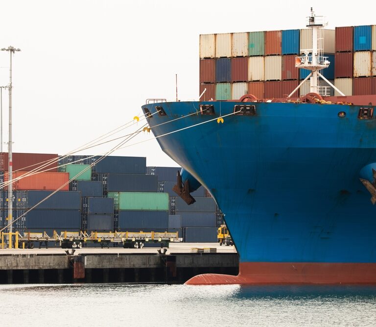 The bow of a large blue cargo ship docked at a port, with stacked colorful shipping containers in the background and mooring lines securing the vessel.