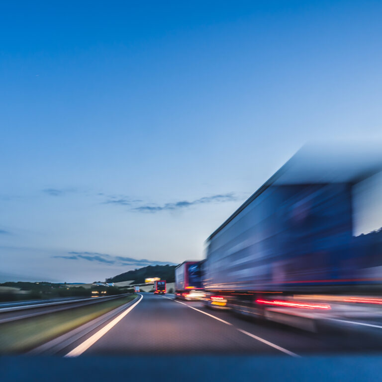 Blurred image of a truck driving fast on a highway at dusk, with a clear sky and distant hills in the background. The motion blur suggests high speed and movement.