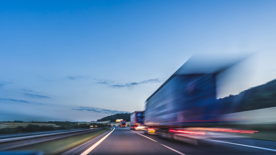 Blurred image of a truck driving fast on a highway at dusk, with a clear sky and distant hills in the background. The motion blur suggests high speed and movement.