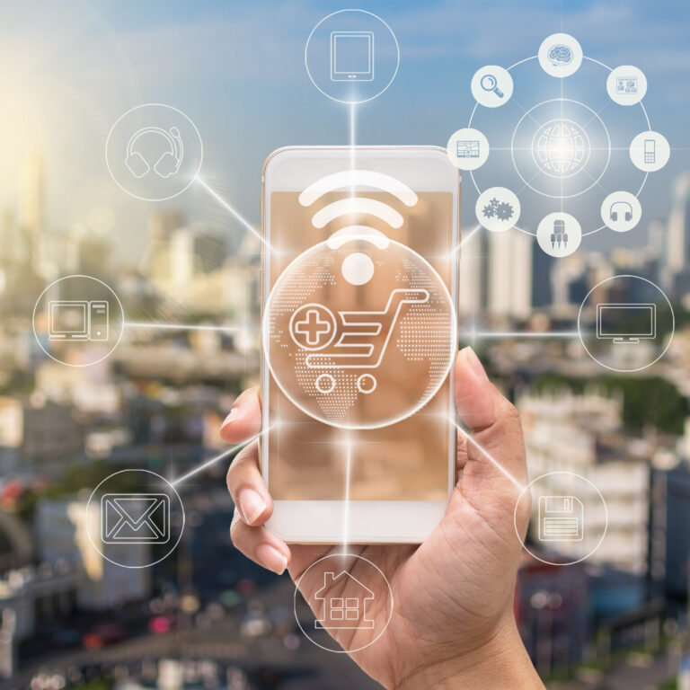A hand holds a smartphone with a glowing shopping cart icon and digital network symbols, representing online shopping, order management systems, and connectivity against a blurred cityscape background.