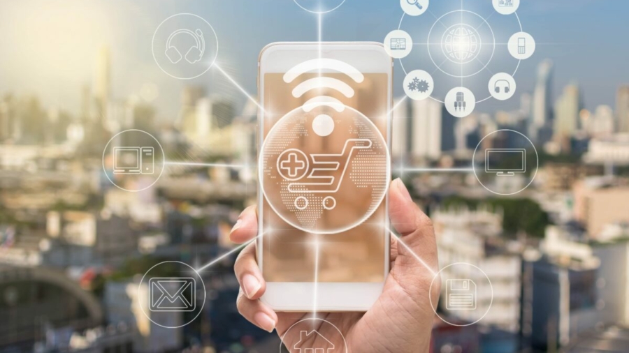 A hand holds a smartphone with a glowing shopping cart icon and digital network symbols, representing online shopping, order management systems, and connectivity against a blurred cityscape background.