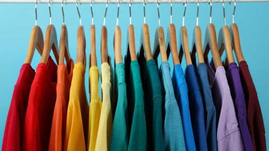 A row of T-shirts in various colors arranged in a rainbow spectrum hangs on wooden hangers from a white clothing rack against a light blue background.