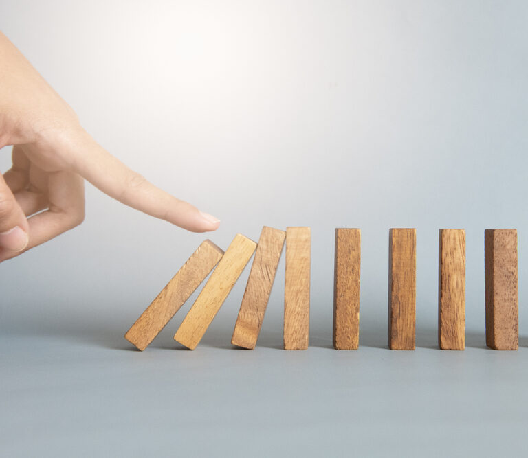 A hand is about to push the first of several standing wooden dominoes, illustrating a bullwhip effect chain reaction. The dominoes are arranged in a line on a smooth surface against a plain background.