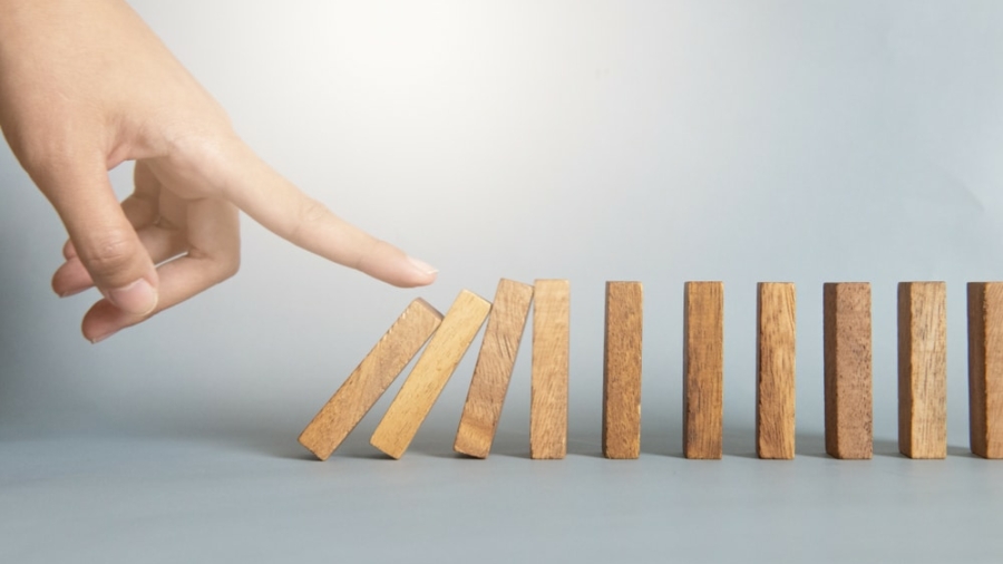 A hand is about to push the first of several standing wooden dominoes, illustrating a bullwhip effect chain reaction. The dominoes are arranged in a line on a smooth surface against a plain background.