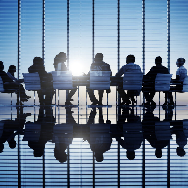 Silhouettes of people sitting around a conference table in a modern office with large windows and blinds, sunlight shining through, and reflections visible on the polished floor.