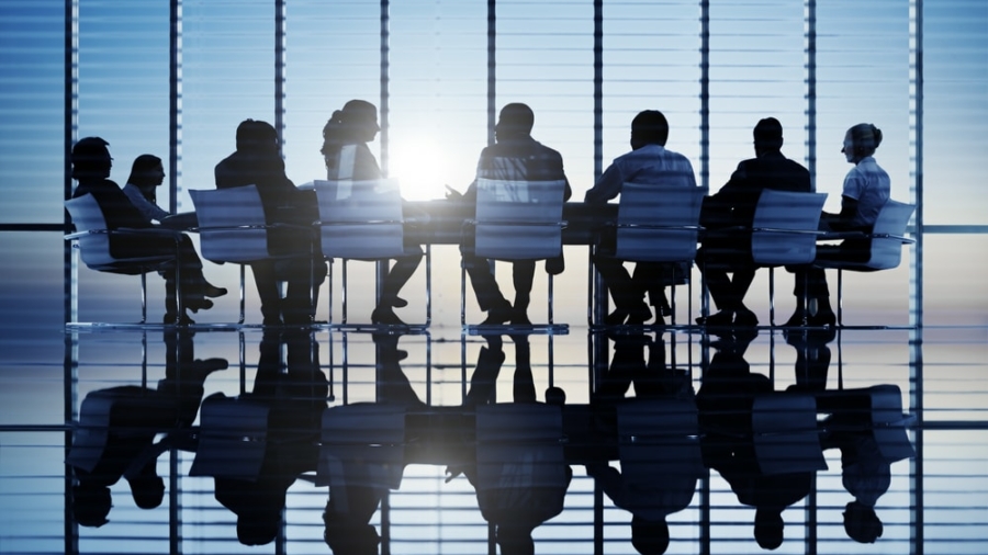 Silhouettes of people sitting around a conference table in a modern office with large windows and blinds, sunlight shining through, and reflections visible on the polished floor.