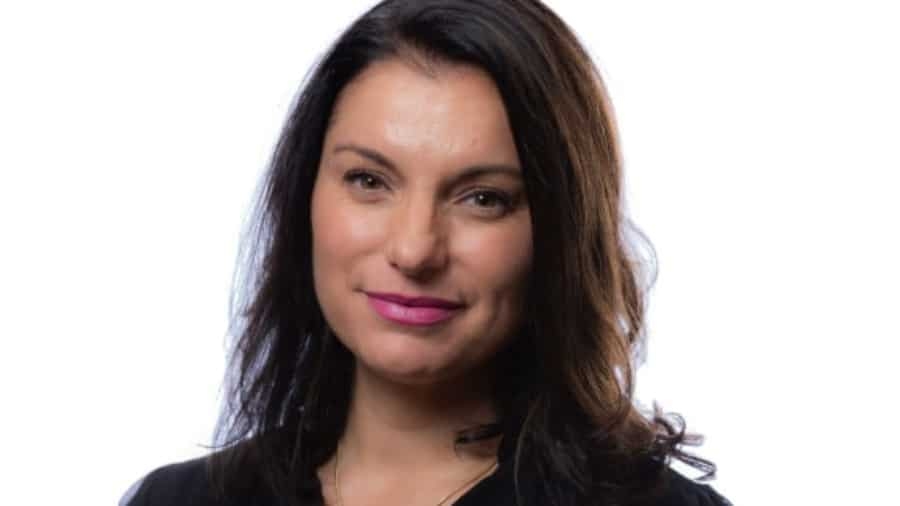 A woman with long dark hair and a slight smile is wearing a black top, posing against a plain white background.