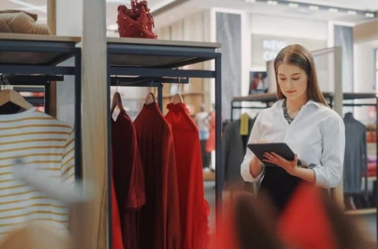 A woman in a white blouse stands in a clothing store, using a tablet. She is surrounded by racks of red dresses and other garments, appearing focused on her work.