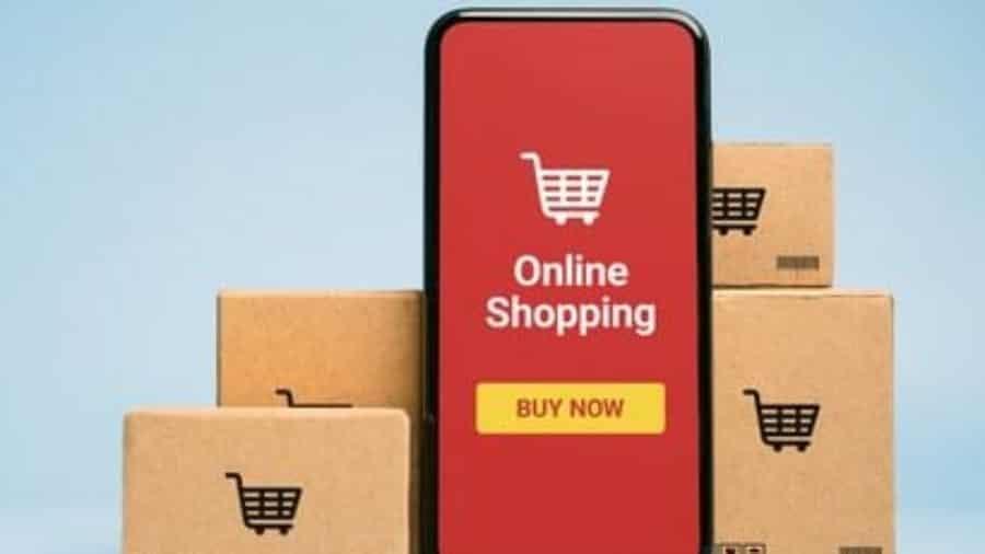 A smartphone displaying an Online Shopping screen with a shopping cart icon and Buy Now button is placed in front of stacked cardboard boxes with shopping cart symbols.