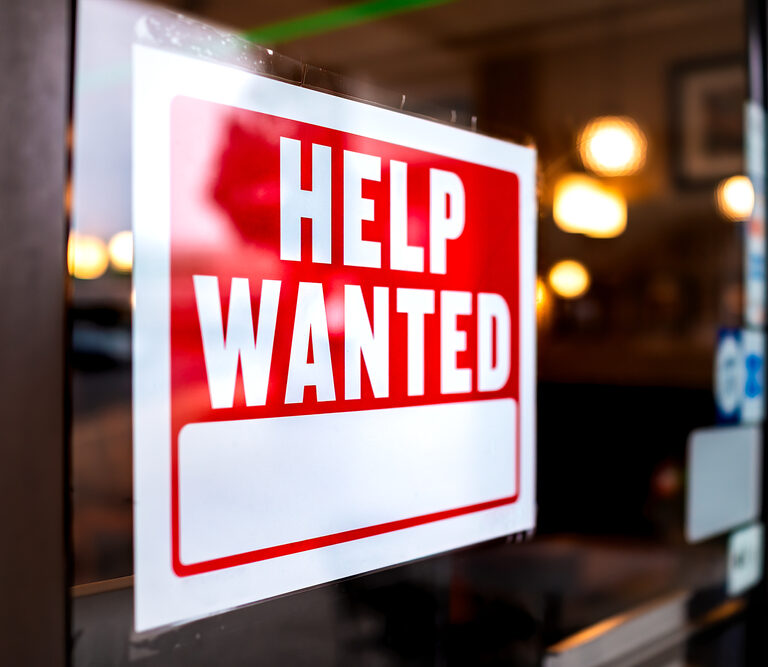 A red and white HELP WANTED sign is displayed in a window, indicating that the business is hiring. The background shows a warmly lit interior with blurred lights.
