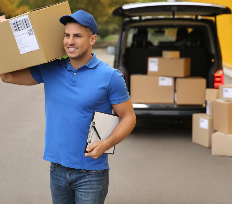 A delivery worker in a blue uniform and cap carries a large cardboard box on his shoulder and holds a clipboard. Several more boxes are stacked near an open van in the background.
