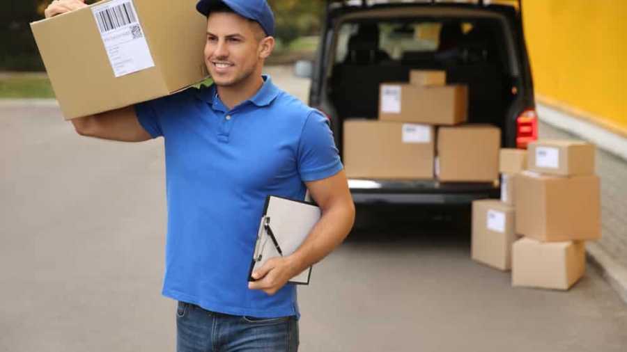 A delivery worker in a blue uniform and cap carries a large cardboard box on his shoulder and holds a clipboard. Several more boxes are stacked near an open van in the background.