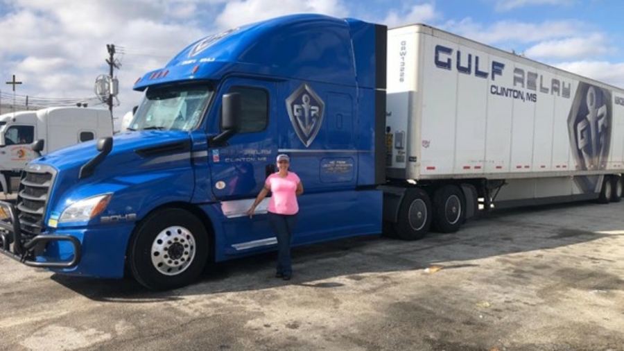 A person in a pink shirt and hat stands next to a large blue Gulf Relay semi-truck and trailer in a parking lot under a partly cloudy sky.