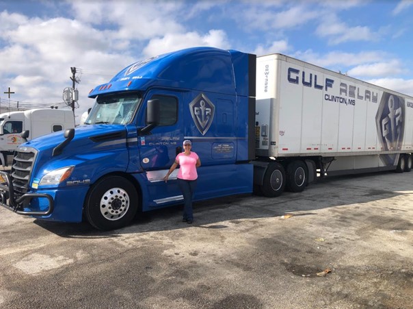 A person in a pink shirt and hat stands next to a large blue Gulf Relay semi-truck and trailer in a parking lot under a partly cloudy sky.