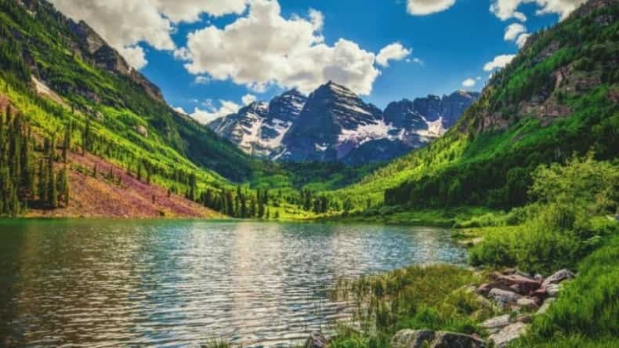 A serene alpine lake surrounded by lush green hills and trees, with snow-capped mountains rising dramatically in the background under a partly cloudy blue sky.