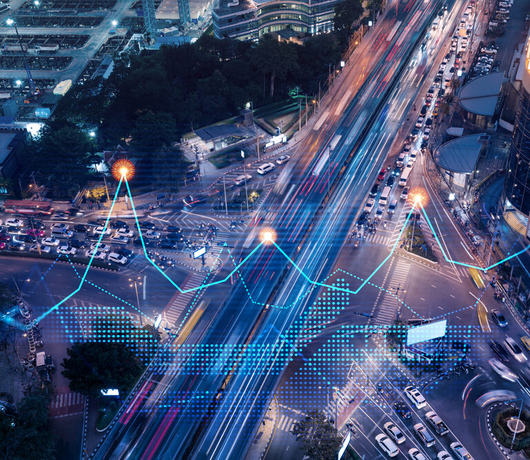 Aerial view of a busy city intersection at night with light trails from traffic. Overlaid graphics show data points, lines, and digital grids, representing data analysis or smart city technology.
