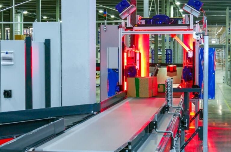 Conveyor belt system in a warehouse with packages being scanned under red lights. The automated machinery and shelving units are visible in the background, indicating a high-tech logistics or sorting facility.