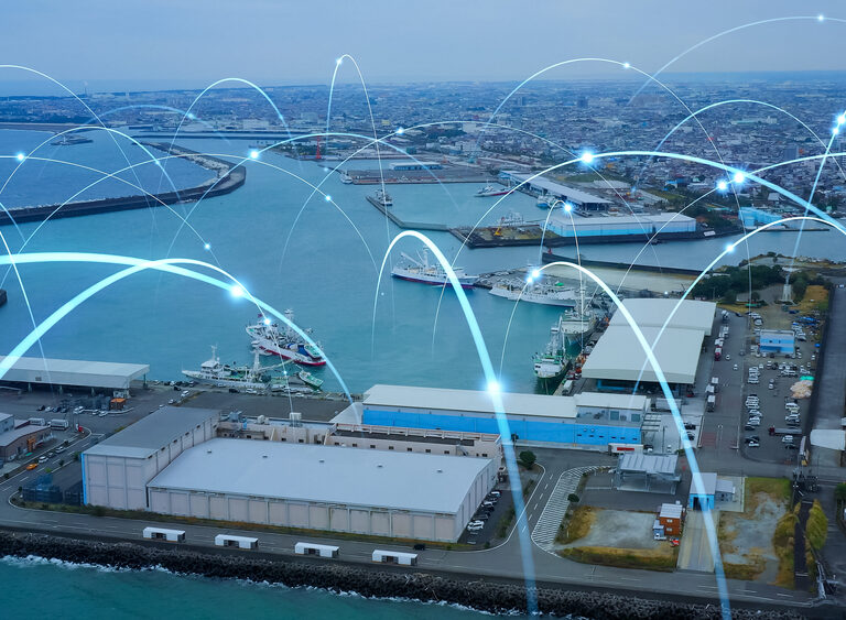 Aerial view of a port city with shipping docks and warehouses, overlaid with digital lines and points representing data communication or a responsive supply chain network.