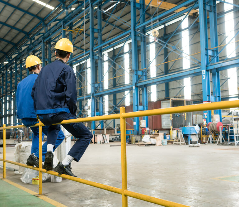 Two workers wearing yellow helmets and blue uniforms sit on a yellow metal railing inside a large, spacious industrial warehouse with equipment and supplies visible in the background.