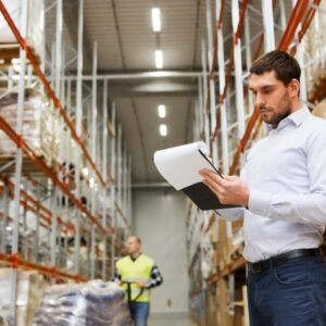 A man in a white shirt stands in a warehouse, holding and reviewing a clipboard. Shelves filled with boxes line the space, and another person in a yellow vest is visible in the background.