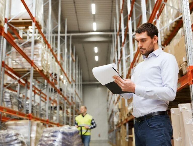 A man in a white shirt stands in a warehouse, holding and reviewing a clipboard. Shelves filled with boxes line the space, and another person in a yellow vest is visible in the background.