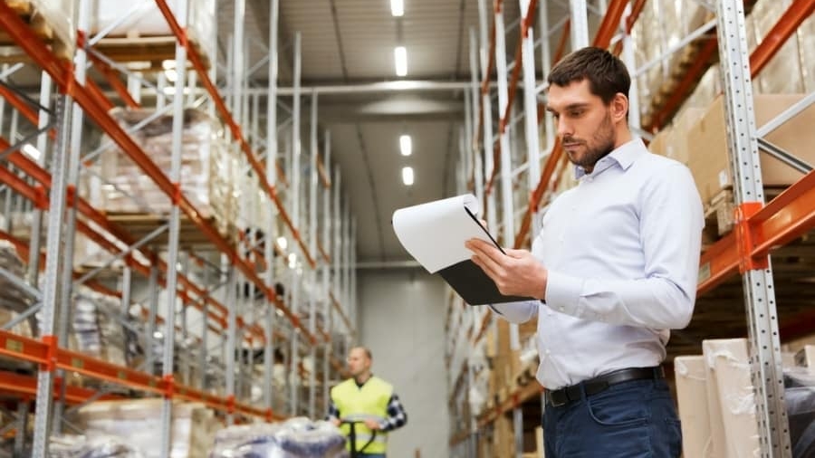 A man in a white shirt stands in a warehouse, holding and reviewing a clipboard. Shelves filled with boxes line the space, and another person in a yellow vest is visible in the background.