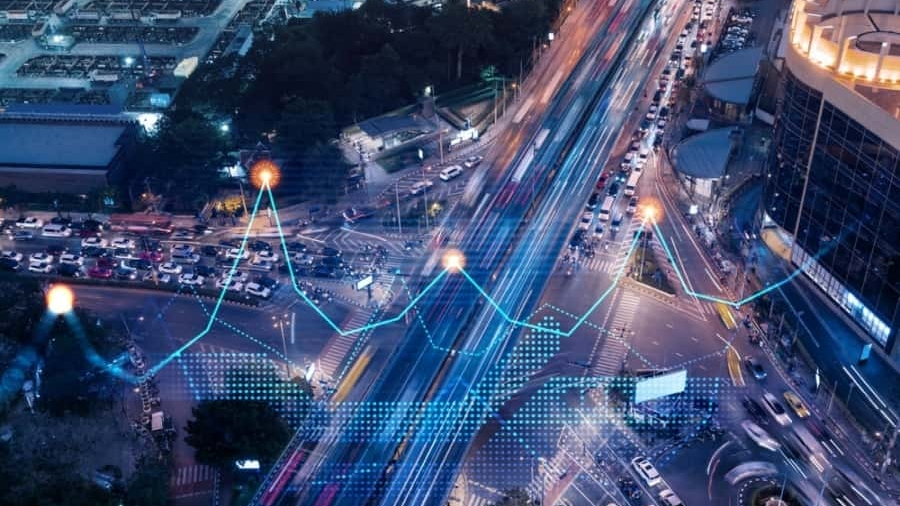 Aerial view of a busy city intersection at night with light trails from traffic and digital lines and data points overlayed, representing smart city technology and data analysis.