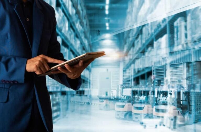 A person in a blue suit uses a digital tablet in a warehouse with tall shelves and automated machinery, suggesting technology-driven inventory or logistics management.