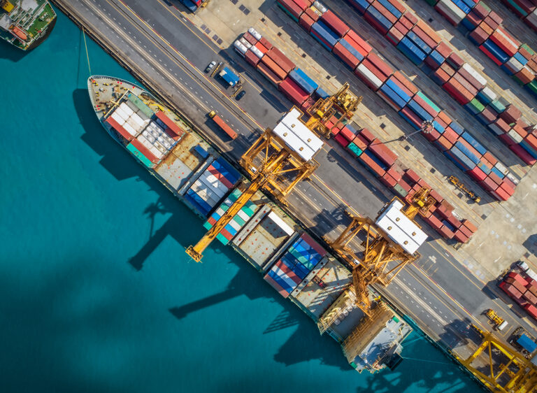 Aerial view of a shipping port with large cargo ships docked, yellow cranes loading containers, and stacks of colorful shipping containers—highlighting the vital role ports play in global supply chains—lined up along the dock next to bright blue water.