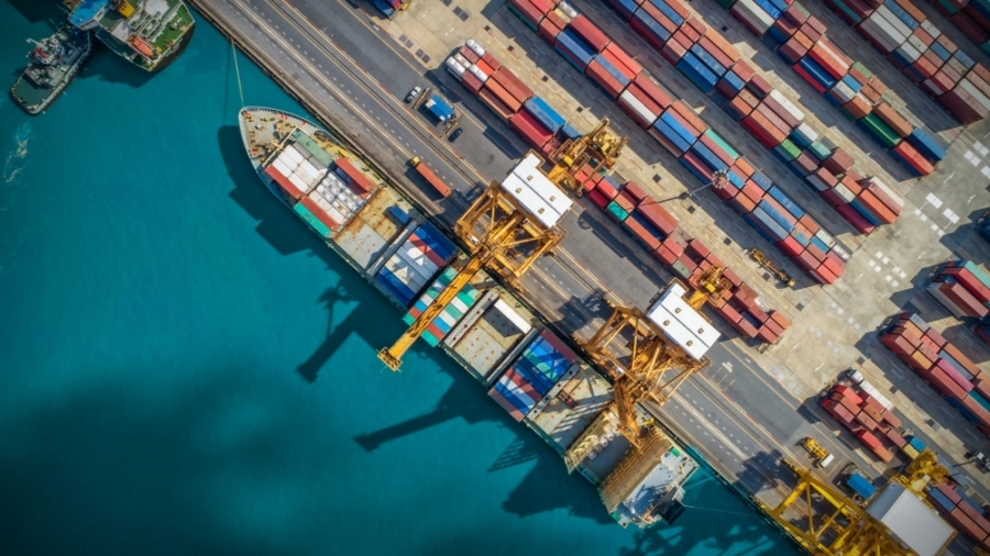 Aerial view of a shipping port with large cargo ships docked, yellow cranes loading containers, and stacks of colorful shipping containers—highlighting the vital role ports play in global supply chains—lined up along the dock next to bright blue water.