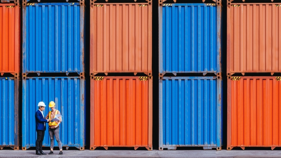 Two people wearing hard hats, one in a suit and one in a reflective vest, stand in front of stacked orange and blue shipping containers, having a discussion.