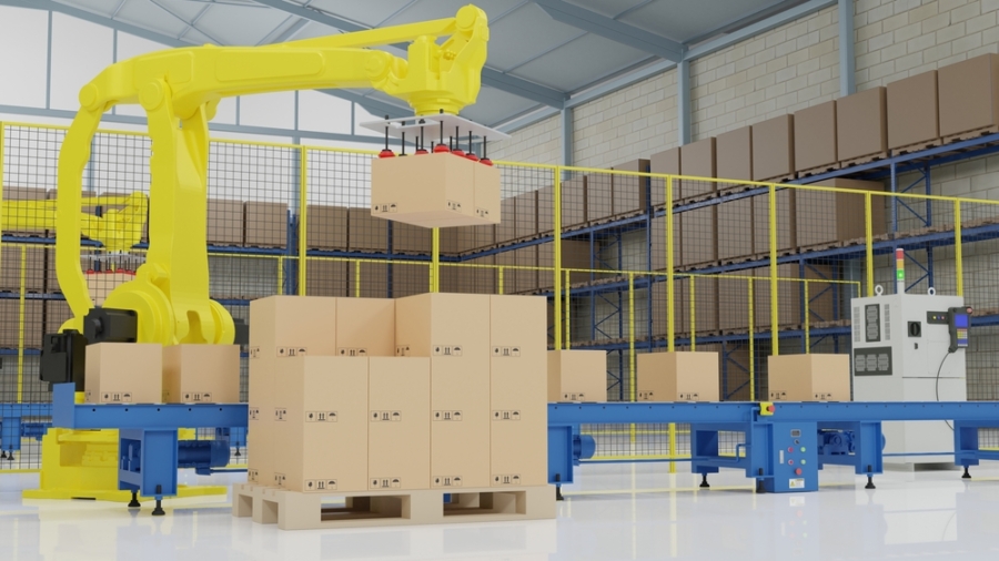 A yellow robotic arm performs automated palletization, moving cardboard boxes onto a conveyor belt in a warehouse filled with shelves of boxes and industrial equipment.