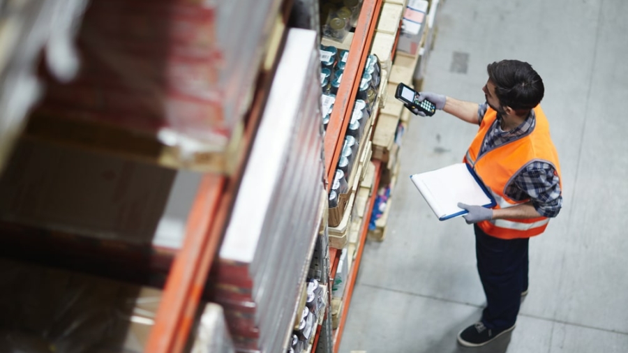 A warehouse worker in an orange safety vest scans items on a high shelf with a handheld scanner while holding a clipboard, standing in an aisle with organized shelves of goods.
