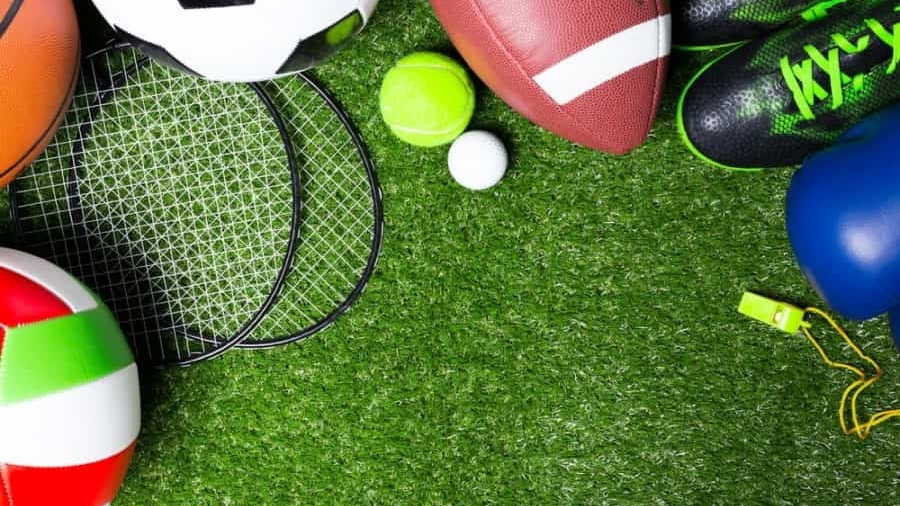 Various sports equipment, including soccer ball, football, volleyball, tennis ball, baseball, badminton rackets, shoes, whistle, and boxing glove, arranged on green artificial grass.