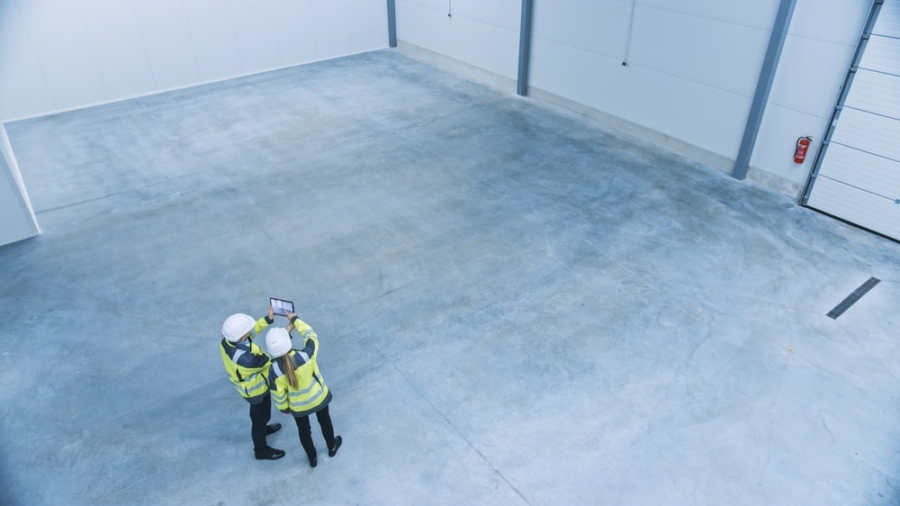Two people in high-visibility jackets and hard hats stand in an empty warehouse, looking at a tablet as they discuss warehouse planning. The warehouse features a concrete floor and white walls.