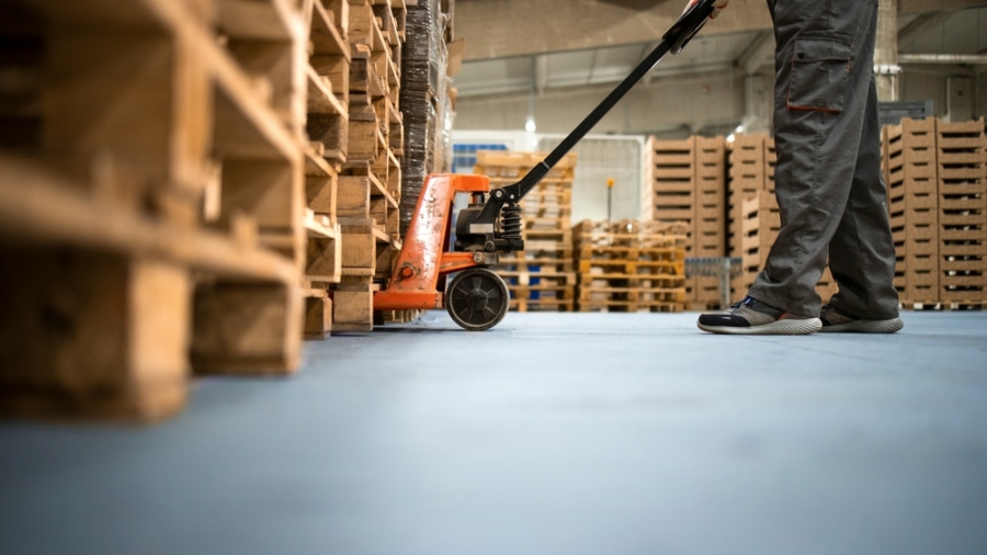 A worker in gray pants and safety shoes uses an orange pallet jack to move wooden pallets inside a warehouse, with stacks of pallets visible in the background.