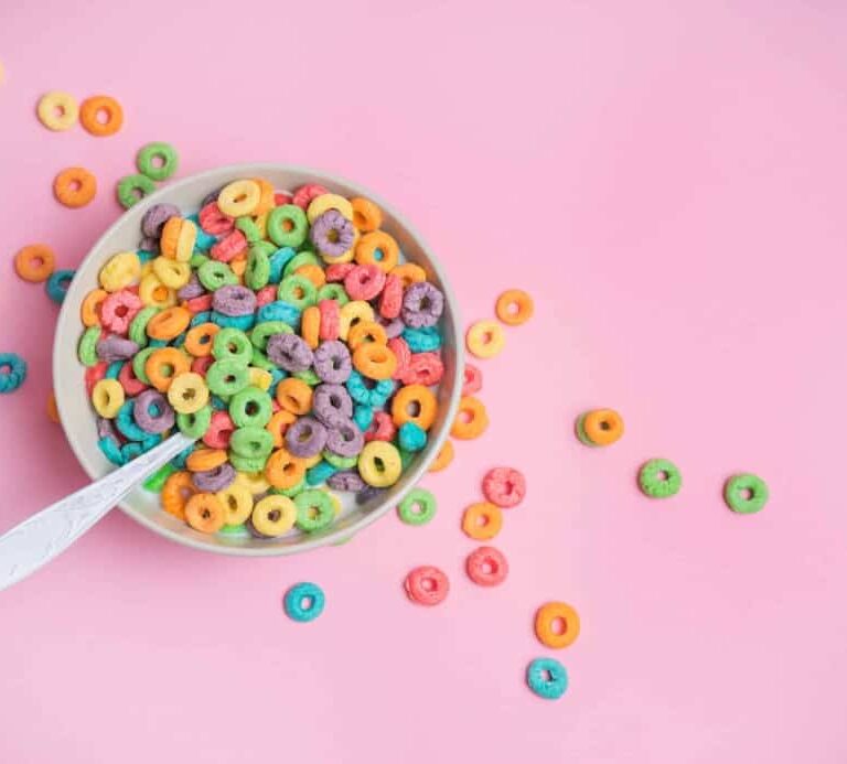 A bowl of colorful, round fruit-flavored cereal with a spoon sits on a pink background, with some cereal pieces scattered around the bowl.