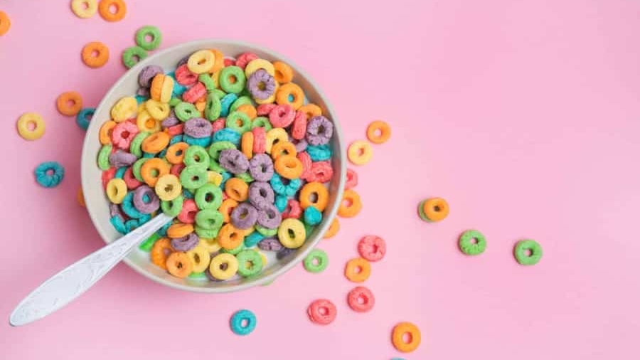 A bowl of colorful, round fruit-flavored cereal with a spoon sits on a pink background, with some cereal pieces scattered around the bowl.