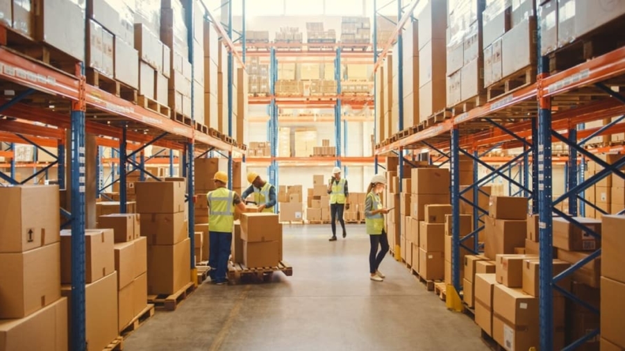 Warehouse workers in safety vests organize and move cardboard boxes on pallets between tall shelves filled with packages in a brightly lit, spacious storage facility.