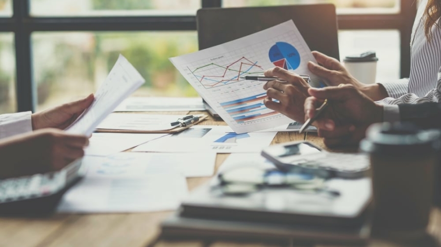 Three people sit at a table reviewing documents with charts and graphs. Laptops, papers, and coffee cups are on the table, suggesting a business meeting or collaborative discussion. Sunlight streams in through large windows.