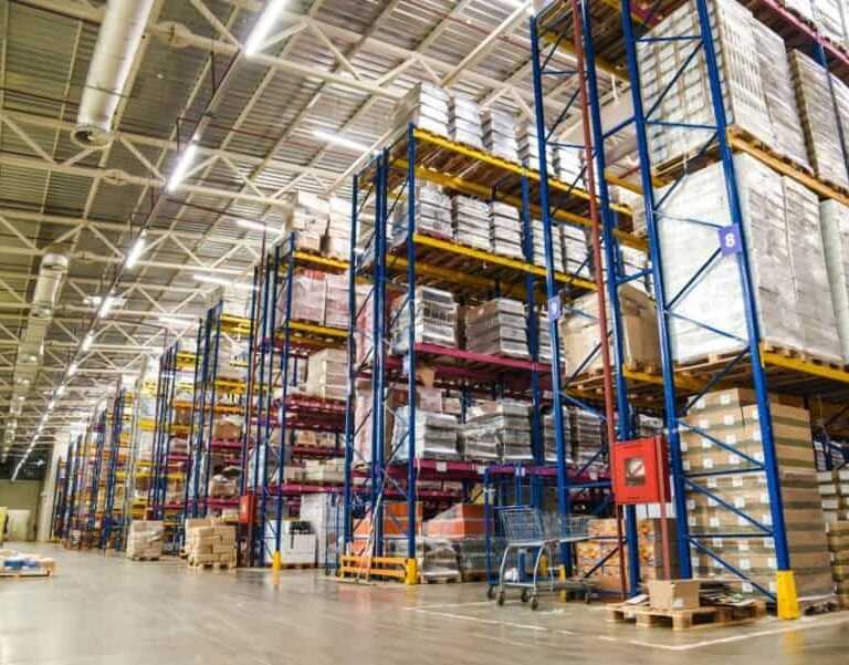Large warehouse interior with tall blue and yellow metal racks filled with stacked pallets and boxes. The concrete floor is clear, and the area is well-lit with overhead lights. Some carts and equipment are visible.