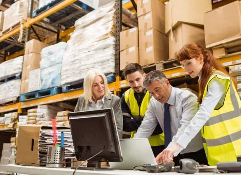 Four staff members, wearing safety vests, collaborate at a desk in a warehouse filled with shelves of boxes and supplies. Two people look at a computer monitor while two others work with papers and a laptop.